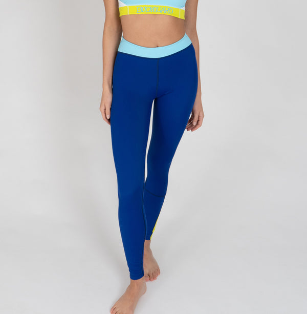 Eine Frau trägt eine dunkelblaue Sportleggings mit einem hellblauen Bund und einem gelben Akzentstreifen mit Bodyguard-Logoam Bein unten