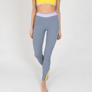 Eine Frau trägt eine graue Sportleggings mit einem lavendel Bund und einem gelben Akzentstreifen mit Bodyguard-Logo am Bein unten.