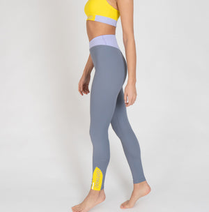 Eine Frau trägt eine graue Sportleggings mit einem lavendel Bund und einem gelben Akzentstreifen mit Bodyguard-Logo am Bein unten.